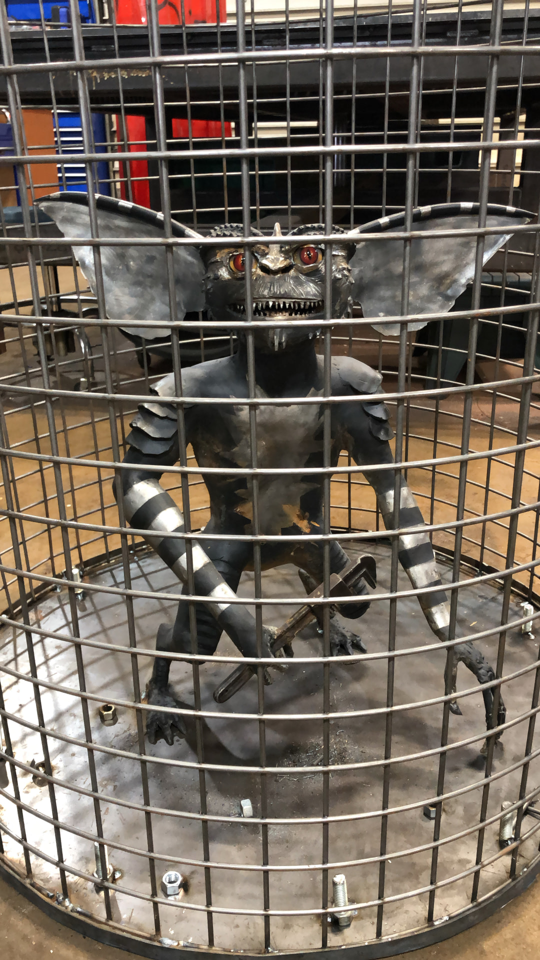Metal creature sculpture with large ears and bared teeth standing inside a round steel wire cage in the workshop